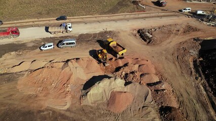 Top-down aerial drone view of a yellow dump truck being loaded with soil by an excavator on a dusty construction site, with rough ground textures, tire tracks, and a worker in a safety vest highlighti
