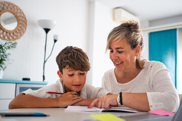 Mother and son are focused on a homework task at a table in a well-lit room. The adult is helping...