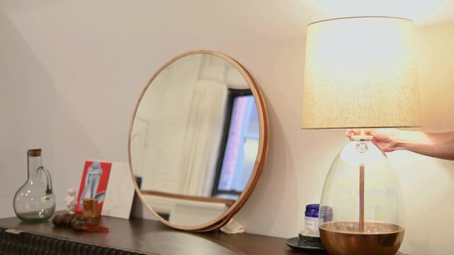 Hand reaching to switch on lamp beside mirror on modern wooden dresser