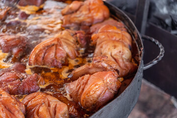 Traditional braised pork knuckles (Schweinshaxe) simmering in a large cauldron at an outdoor food festival or market.Close-up of savory pork shanks being slow-cooked in a rich sauce