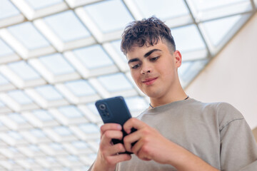 Smiling teenage boy using smartphone in modern building. Happy young man texting on mobile phone. Teen boy in gray t-shirt typing on cellphone. young man with smartphone. Copyspace