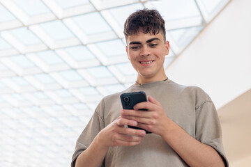 Smiling teenage boy using smartphone in modern building. Happy young man texting on mobile phone....