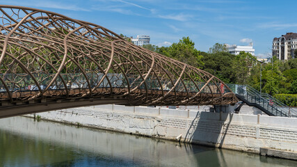 Modern, intricately designed pedestrian bridge arches over calm river Sochi, set against a backdrop of lush greenery and urban buildings under clear blue sky. Sochi, Russia - May 23, 2025