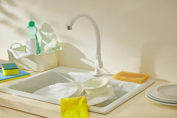 Sink with stacked dishes, sponge, and cleaning products. The yellow cloth adds brightness to a regular kitchen scene prepared for cleaning.