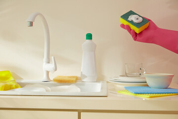 Hands wearing pink gloves with detergent foam on a sponge are preparing to clean the kitchen, rinsing dishes in the sink. Casual photo shoot for dishwashing product advertisement