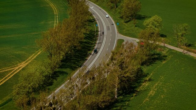 Cars moving along winding paved road through countryside fields. Vehicles driving on curvy asphalt route amid rural landscape. Automobiles traveling through bendy tarmac highway across farmland