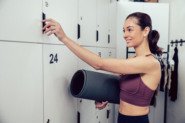 Smiling young woman with a rolled mat opening a locker in the gym