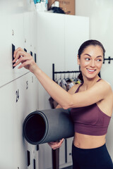 Smiling young woman with a rolled mat opening a locker in the gym