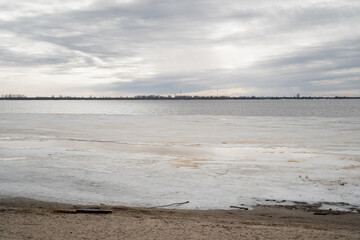 The wide northern river with a sandy bank was partially freed from winter snow and ice on a cloudy day
