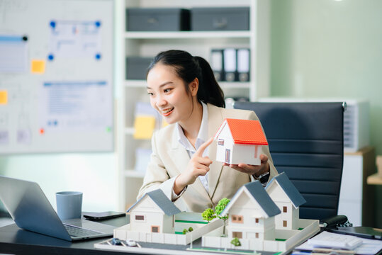 Asian real estate agent in modern office consulting clients, showcasing properties with a laptop, tablet, and house model. - Powered by Adobe