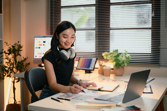 Graphic designer woman is sitting at a desk with a laptop and a notebook. She is wearing headphones and smiling