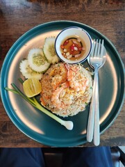 Shrimp fried rice on a dark green plate, garnished with cucumber and spring onions, accompanied by a small bowl of prik nam pla (chili fish sauce).