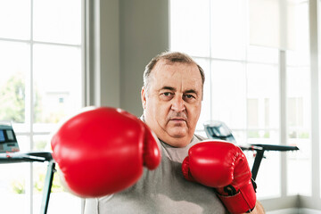 Confident overweight senior man wearing red boxing gloves, throwing a punch at the gym. Promotes active aging, fitness, and strength through boxing as part of a healthy, determined lifestyle.
