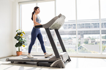 Confident young woman walking on a treadmill in a bright indoor gym with large windows, embracing wellness, fitness, and a healthy lifestyle through daily cardio and mindful exercise routines.