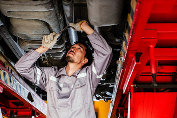 Auto mechanic in gray uniform tightening components under a raised car in a repair shop, performing hands-on maintenance with a wrench during routine vehicle inspection and mechanical servicing.