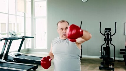 Confident overweight senior man wearing red boxing gloves, throwing a punch at the gym. Promotes active aging, fitness, and strength through boxing as part of a healthy, determined lifestyle. - Powered by Adobe