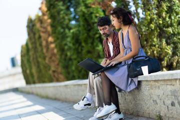 Two young adults collaborating outdoors on a laptop, seated on a stone ledge in a park, surrounded by greenery and sunlight