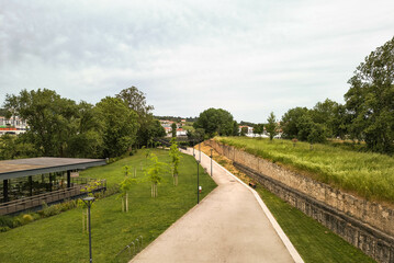 A picturesque walkway winds through a vibrant park, framed by lush greenery, old stone walls, and a modern glass building. Tranquil scenery evokes peace, inviting contemplation.