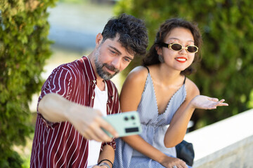 Joyful young couple taking a selfie outdoors on a sunny day, capturing a happy moment together with a smartphone, wearing casual summer outfits and sunglasses
