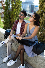 Casual young couple outdoors in an urban park sitting on a stone bench with a laptop in stylish outfits and sunglasses enjoying a sunny day with greenery in the background