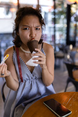 Young woman enjoying a chocolate ice cream cone in a cozy cafe with stylish summer dress and playful expression