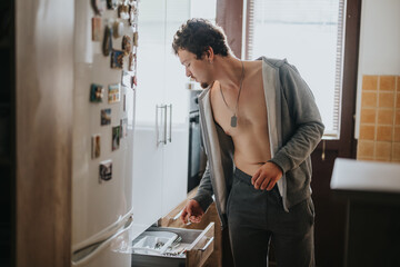 A young man dressed casually searches a kitchen drawer, surrounded by everyday objects, in a well-lit domestic environment.