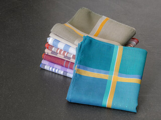 A neatly folded stack of colorful striped and plaid handkerchiefs rests on a dark surface, with one prominent teal-blue handkerchief in the foreground.