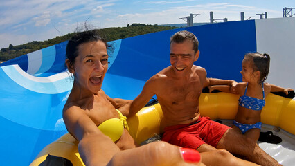 Medium shot captures family enjoying water slide at aqua park. Woman in yellow bikini, and young girl in blue polka dot bikini pose for selfie, showcasing their excitement and fun in summer.