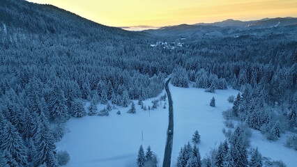 An aerial view of a winding road through a snowy forest, with a car driving along the path. The scene evokes a sense of winter travel and alpine adventure.