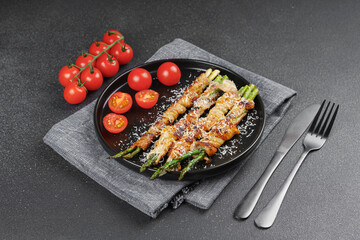 Grilled asparagus wrapped in crispy bacon sprinkled with parmesan cheese served on plate with fresh cherry tomatoes on black background. Gourmet dish, cooking recipe, cuisine, or restaurant menu
