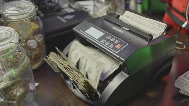 A stack of cash runs through a money counter in a studio setting, surrounded by glass jars and creative clutter &mdash; a glimpse into the hustle workflow.