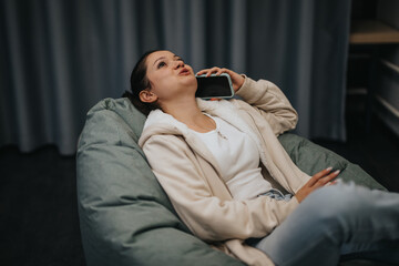 Fototapeta premium Casual scene of a young woman reclining comfortably on a bean bag while talking on her phone, exuding relaxation and contentment in a cozy indoor setting.