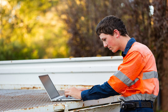 Young trainee tradie apprentice working on laptop in back of ute in high-vis workwear