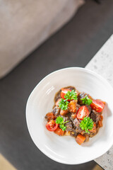 A vibrant dish in a white bowl, featuring chunks of meat, diced carrots, cherry tomatoes, and fresh parsley garnish. Placed on a countertop with a blurred background, it exudes a appetizing appeal.