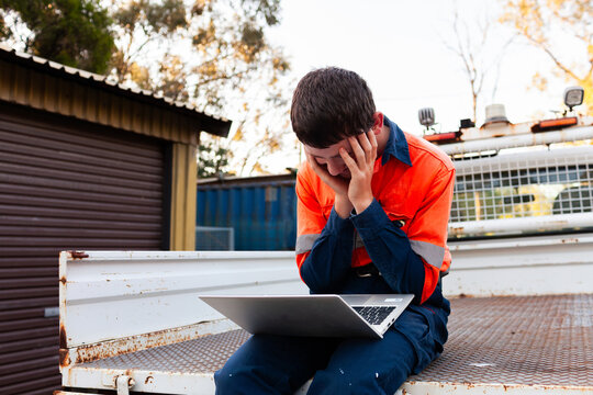 Young stressed trade apprentice working on laptop in back of ute in high-vis workwear
