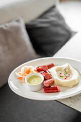 A plated meal featuring sliced sausages, mashed potatoes with garnish, green sauce, and shredded vegetables. The dish is served on a white plate with a blurred background.