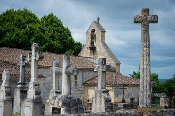 A cemetery filled with ornate stone crosses stands adjacent to a historic church in Saint-Laurent-des-Combes, surrounded by lush greenery under a dramatic sky.
