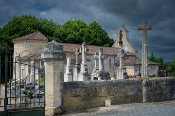 A cemetery filled with ornate stone crosses stands adjacent to a historic church in Saint-Laurent-des-Combes, surrounded by lush greenery under a dramatic sky.