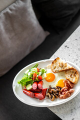 A hearty breakfast plate with sunny-side-up eggs, crispy bacon, grilled bread, sausages, mushrooms, and a fresh salad of lettuce, cherry tomatoes, and cucumber, served on a speckled countertop