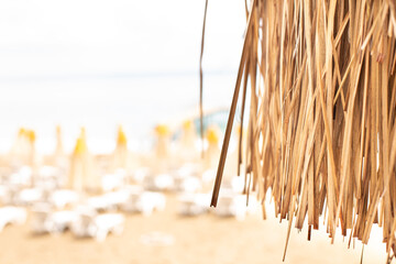 Thatched roof with tropical beach with sand and deck chairs on the sea shore in background.