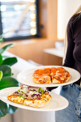 A person holds two plates of pizza—one with pepperoni and the other with assorted toppings, including greens and cheese. The blurred background features a window with lights