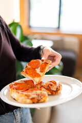 A close-up of a hand lifting a slice of pepperoni pizza from a white plate with two more slices. The melted cheese stretches, enhancing the appeal of this casual dining scene.
