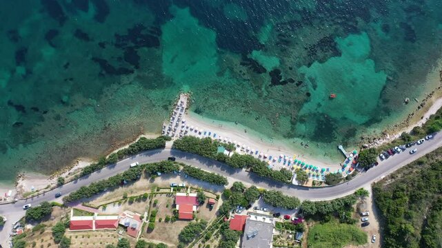 Upward-moving drone shot of Vlora Beach in southern Albanian, revealing turquoise waters, umbrellas lined along the coast, lush greenery, coastal homes, and the scenic clarity of the Ionian shoreline.