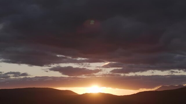Aerial shot of a sunset in the mountains, filmed in 70mm. The dark peaks, golden rays, and a large gray cloud create a dramatic backlit scene as the drone camera slowly descends.