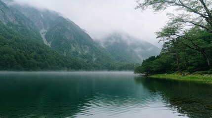 Fototapeta premium Misty morning in Kamikochi valley with fog drifting between tall mountains and calm reflective lake, untouched nature, no people