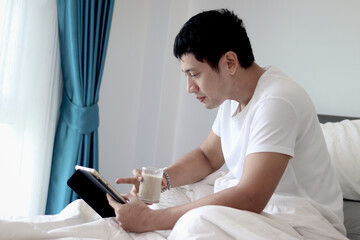 Obraz premium Asian man using tablet while drinking almond milk in bed after waking up in the morning in bedroom. Man working on his tablet during lying down on comfortable bed. Sleepy man surfing internet in bed.