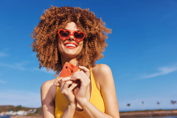 Happy woman in heart shaped sunglasses holding a slice of watermelon at the beach under a clear...