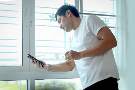 Asian man using smartphone while drinking almond milk, standing by bedroom window in the morning. Man checking his mobile phone during drinking his healthy beverage milk glass after waking up at home.