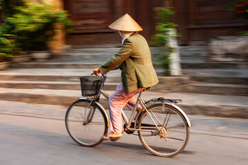 Elderly person riding a bicycle in motion, wearing a traditional conical hat in Vietnam