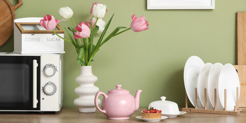 Pink teapot with tasty dessert and beautiful tulip flowers on table near green wall in kitchen